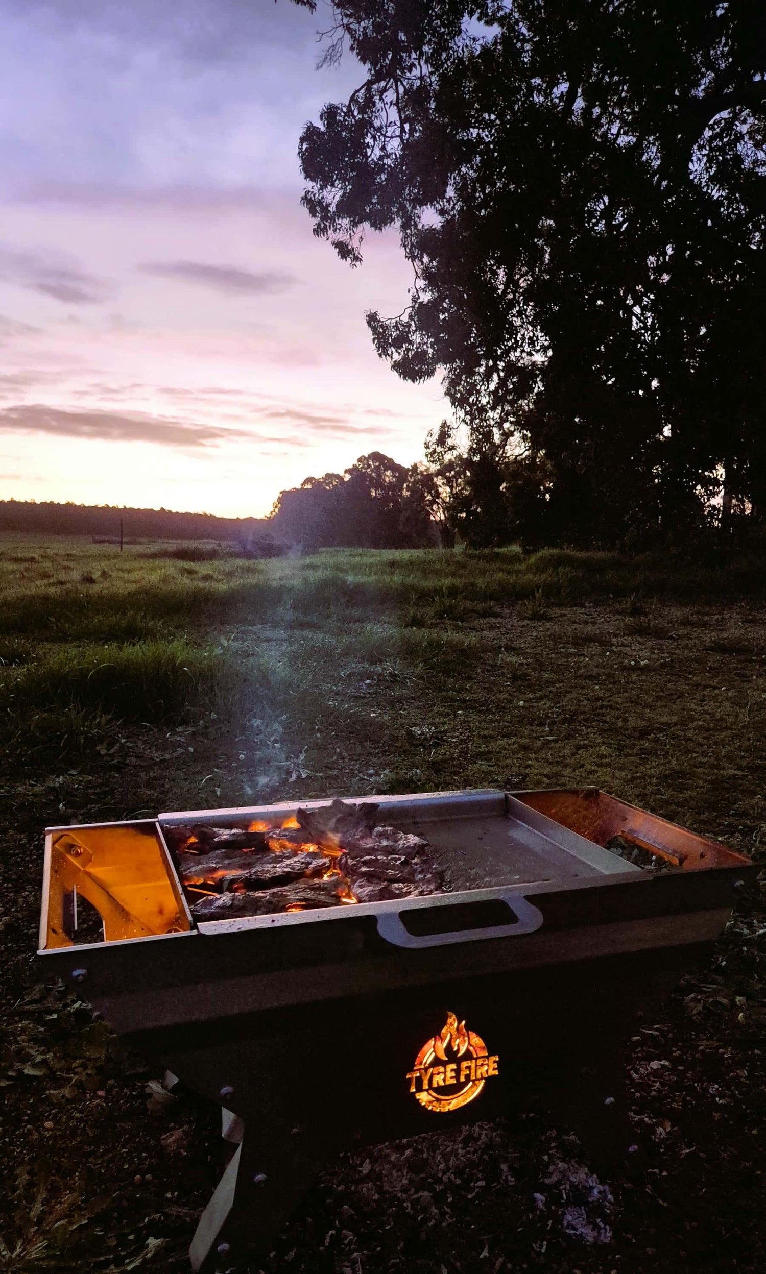 Caravan fire pit at sunset with Tyre Fire logo glowing, stainless steel tyre-mounted BBQ for camping, caravanning and 4WD trips.